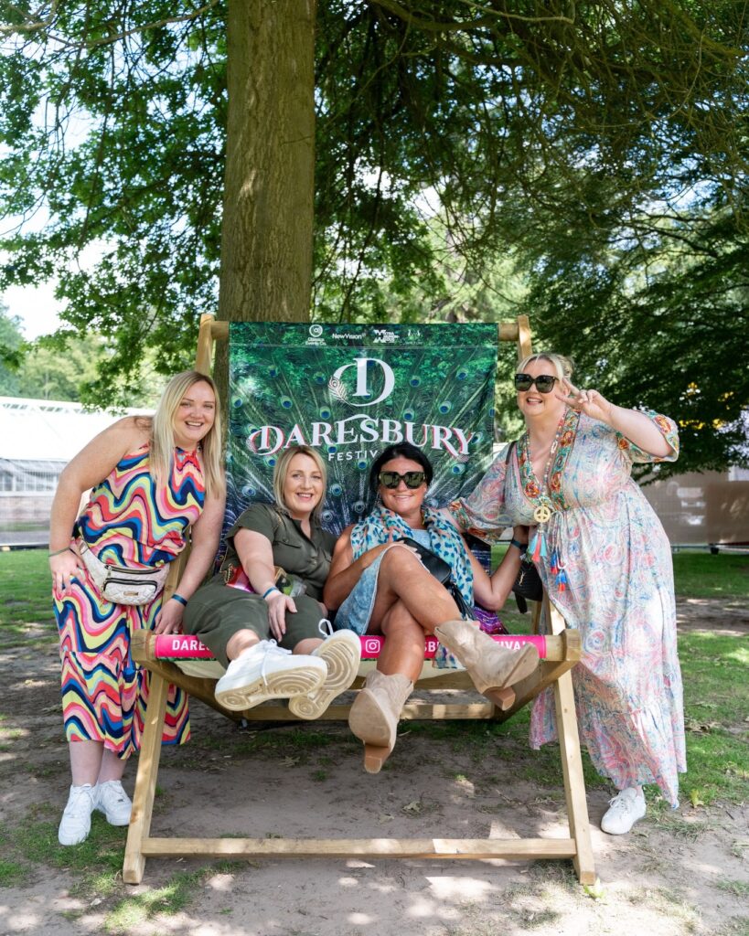 a group of 4 friends sit on a giant Daresbury Festival deckchair, underneath a tree in the shade from the sunshine.