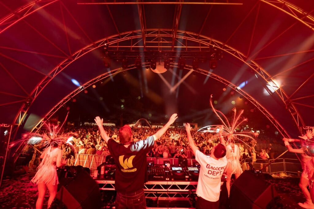 image taken from behind the decks on the stage, showing two DJs with their arms raised looking out to a huge crowd, with two dancers in sparkly outfits either side.