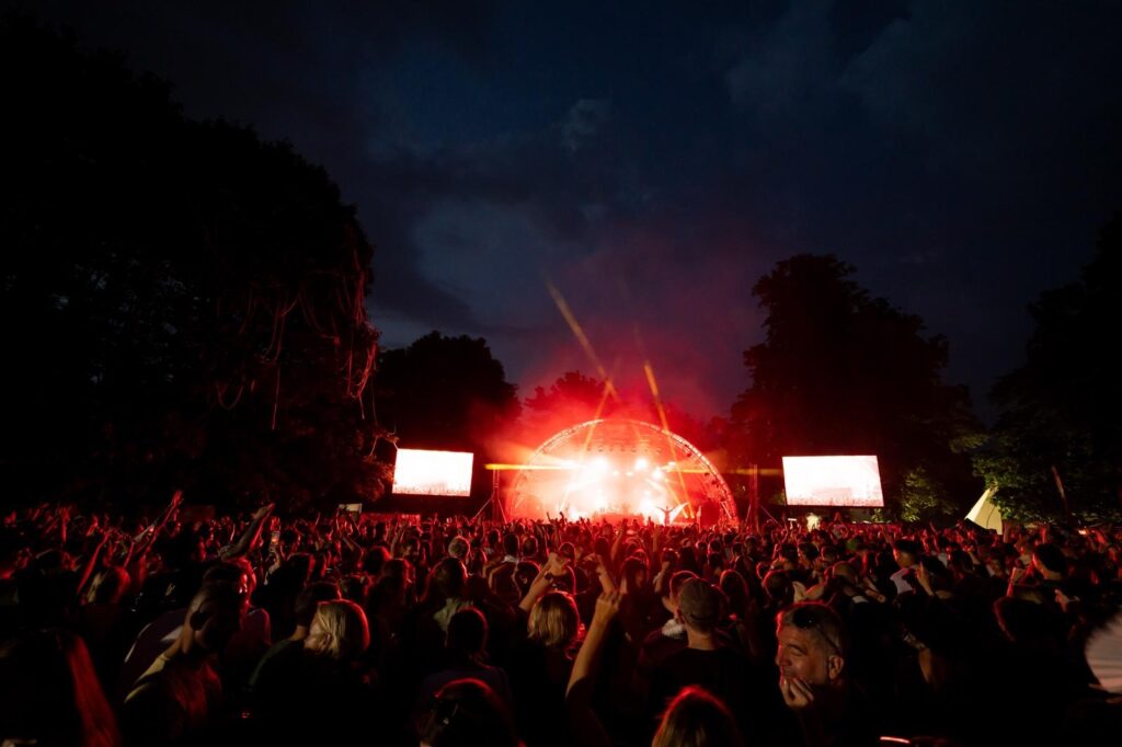 a red light show glows from the stage under a dark sky at night, with a huge crowd watching.