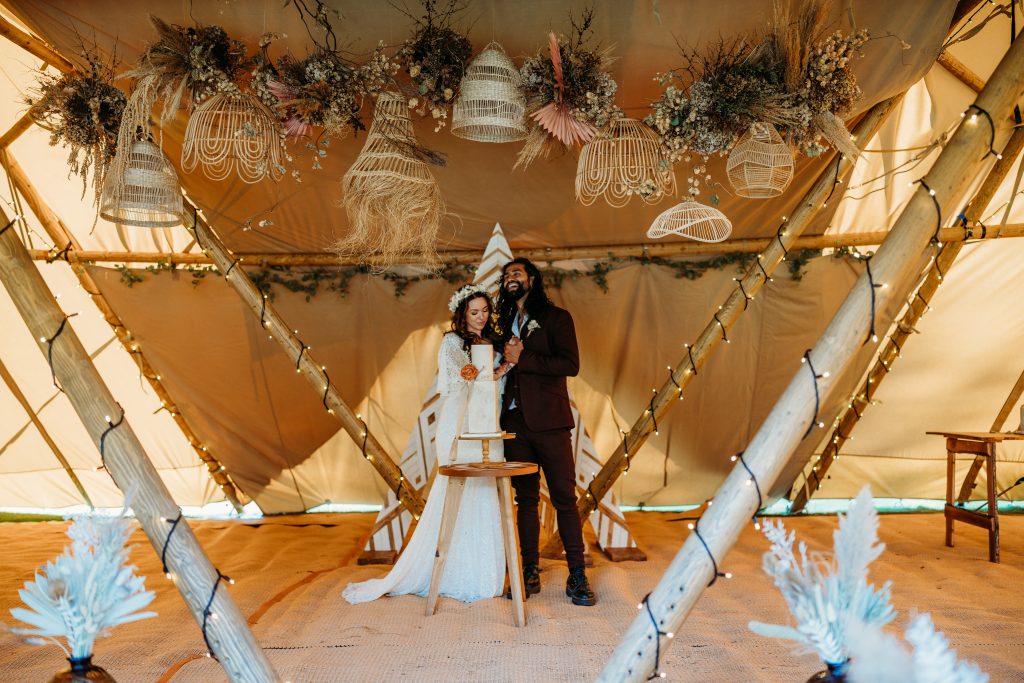 A newly married couple cut their cake inside a tipi, decorated with dried flowers, fairy lights and basket/floral ceiling display.