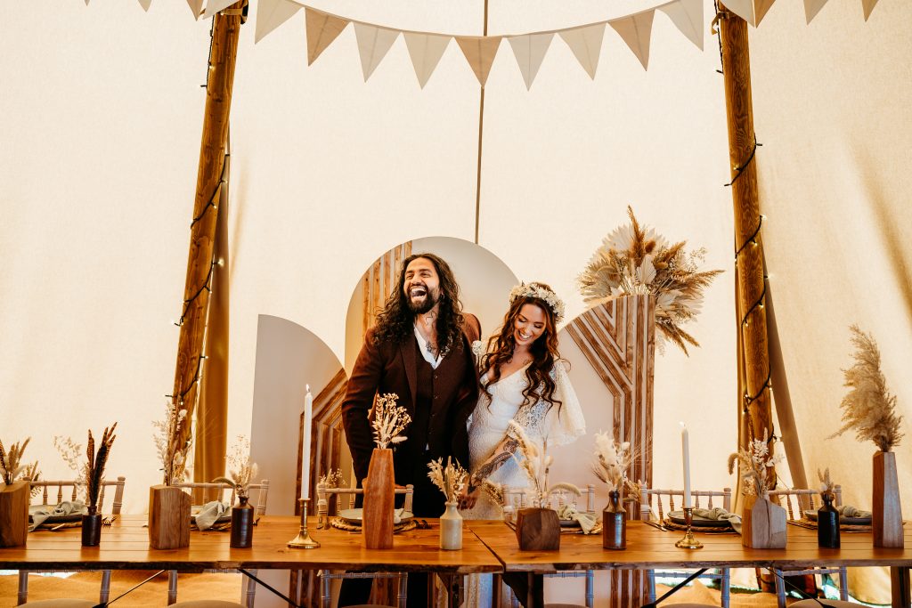 A newly married couple stand inside a tipi, decorated with bunting, vases and dry flowers.