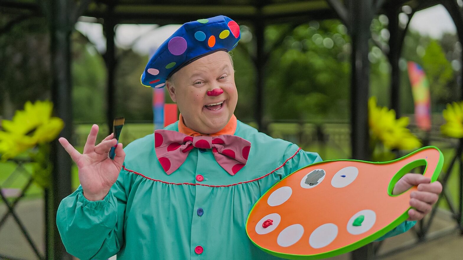 Mr Tumble wearing green shirt with a pink bowtie and a blue beret, whilst holding a paintbrush at the bandstand in Walton.