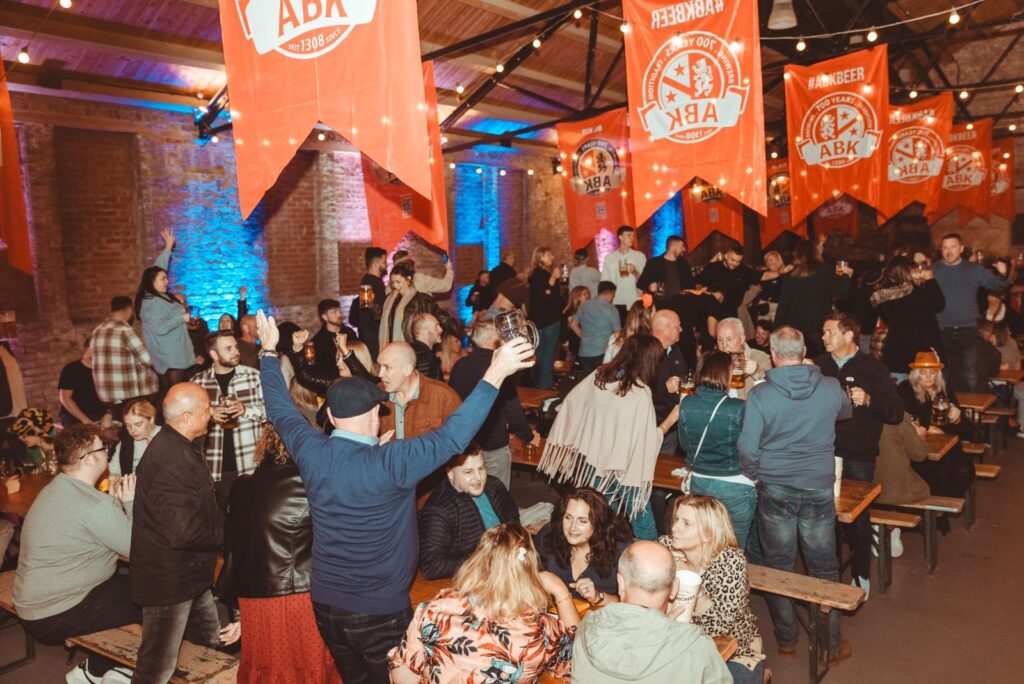 A photo from a Oktoberfest event. a large group of people are standing and seated around/on wooden benches. Red bavarian flags hang from the ceiling of the old riding school, with some people raising their arms in the air enjoying the atmosphere of the event.